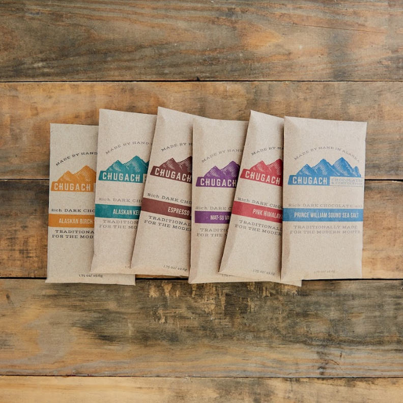 A collection of five Chugach brand chocolate bars, each with different color packaging, displayed on a wooden surface.