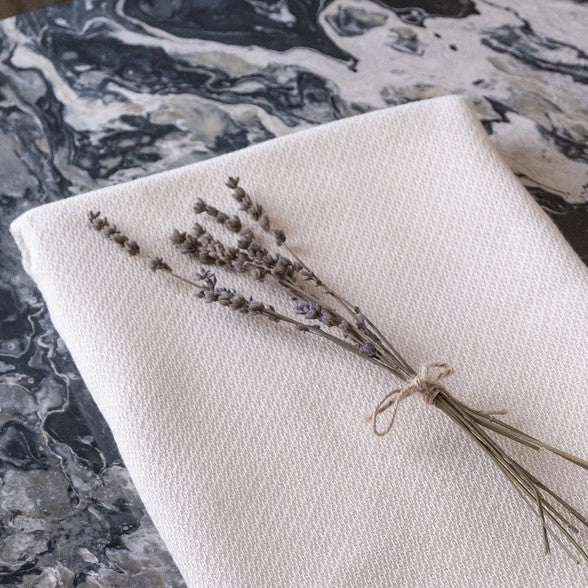 A cream-colored Turkish throw blanket laying flat with a bundle of dried lavender flowers tied with a string on top, placed on a marble surface.