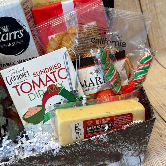 Gift basket with sundried tomato dip mix, cheese, and other snacks on a wooden surface.