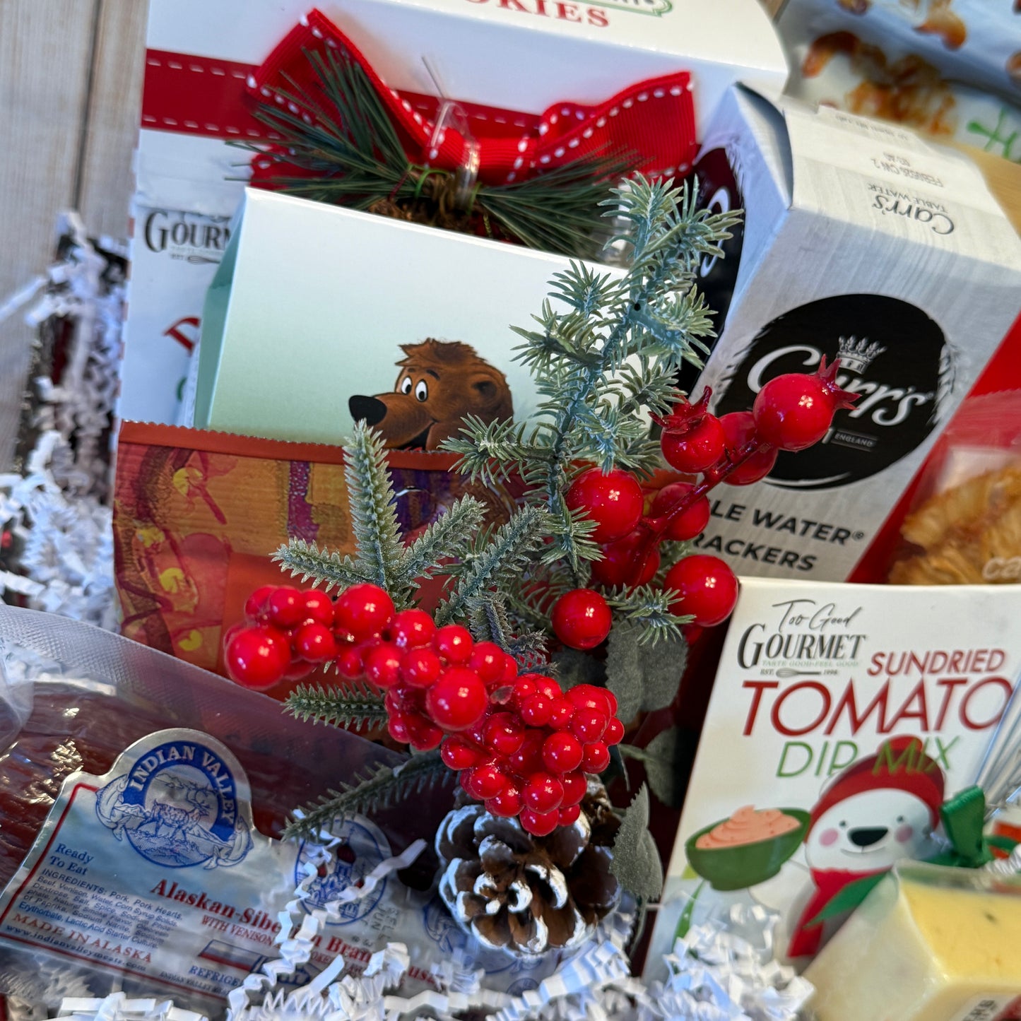 Assorted food items including a box of cookies, a jar of jelly, and a package of sundried tomato dip mix, with decorative elements like berries and pine cones.