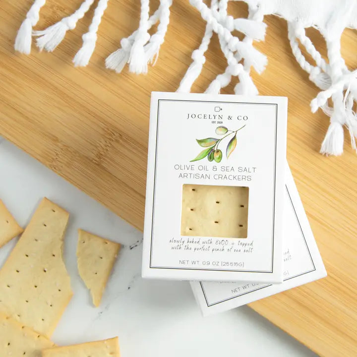 Mini Olive Oil & Sea Salt Crackers by Jocelyn & Co. stacked in boxes, with some crackers displayed nearby, showcasing their artisanal square shape and light seasoning.