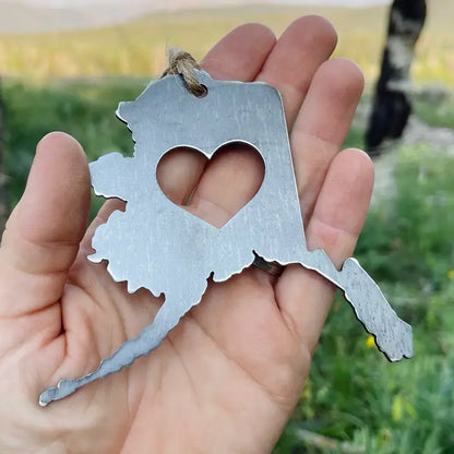 Steel Alaska-shaped keychain with heart cutout held in a hand outdoors.