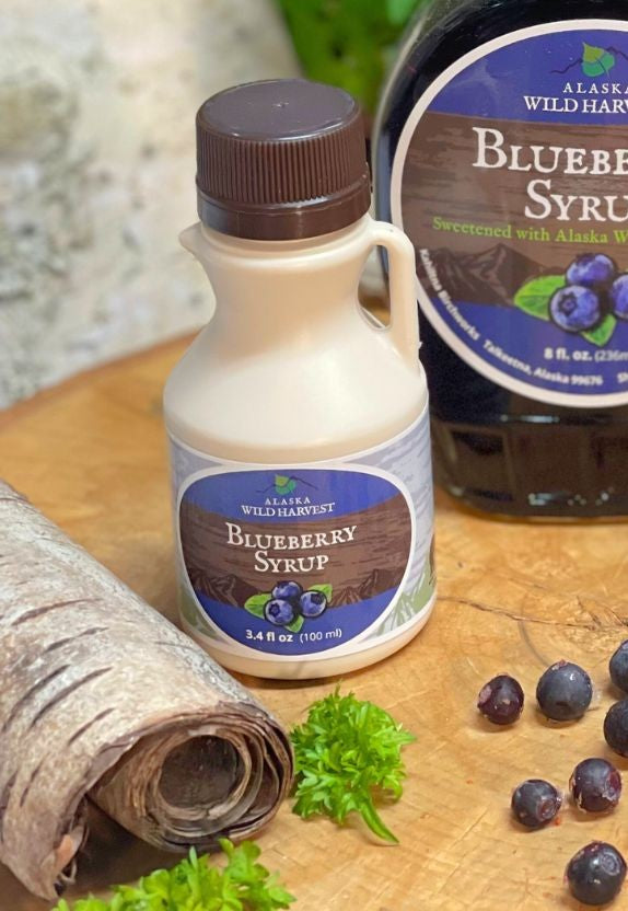 Alaska Wild Harvest Blueberry Syrup bottle on a wooden surface with blueberries and leaves.