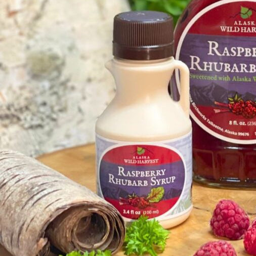 A bottle of Alaska Wild Harvest Raspberry Rhubarb Syrup with a label showing the product name and weight, alongside fresh raspberries and a piece of rhubarb.