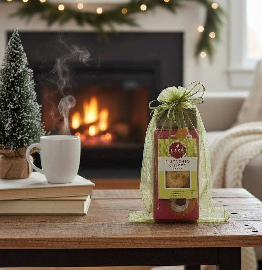Artisan Bites Gift Set with Pistachio Cherry cookies in organza bag, placed on a cozy holiday table near a steaming mug and mini evergreen.