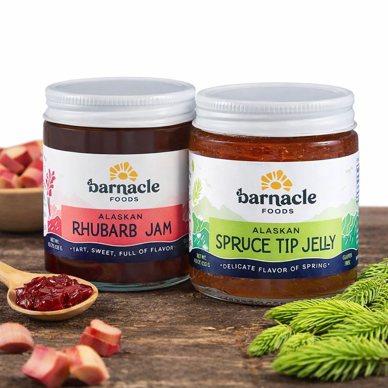 Two jars of Barnacle Foods Alaskan Rhubarb Jam and Spruce Tip Jelly on a wooden surface with rhubarb and spruce tips. 