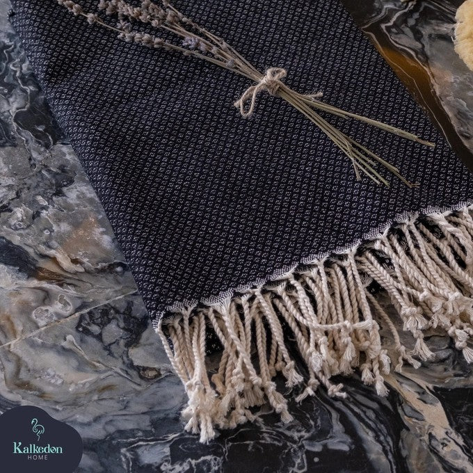 A black-colored Turkish throw blanket laying flat with a bundle of dried lavender flowers tied with a string on top, placed on a marble surface.
