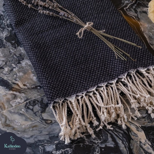 A black-colored Turkish throw blanket laying flat with a bundle of dried lavender flowers tied with a string on top, placed on a marble surface.
