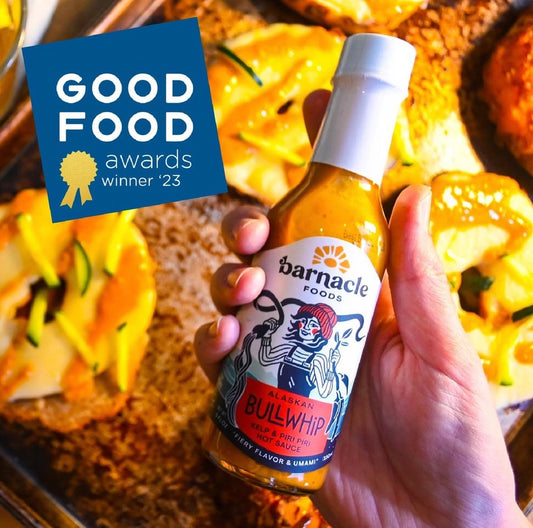 Hand holding a bottle of Barnacle Foods Bullwhip hot sauce with 'Good Food Awards' winner '23' badge in the background.
