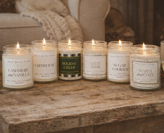 Six lit Candles | 9oz | Long-lasting, Vegan friendly from Sweet Water Décor, showcasing winter scents like Cashmere and Vanilla, and Sugar Cookies, on a rustic surface.