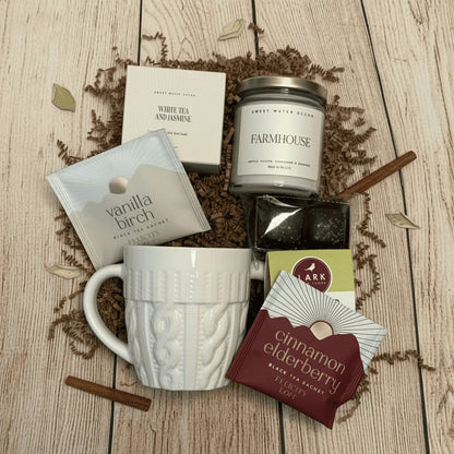 Gift set with a bath bomb, candle, mug, cookie, and tea in a wooden box on a wooden surface.