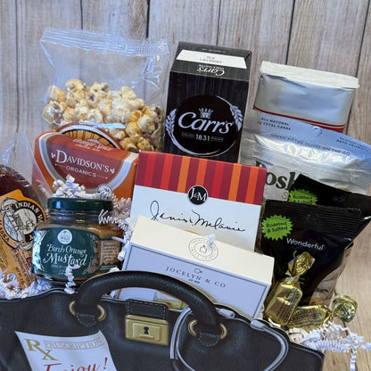 A gift basket with an assortment of food items such as crackers, cheese spread, sausage, mustard, olives, tea, cookies, and pistachio cheese straws, packaged in a decorative black bag with a red ribbon.