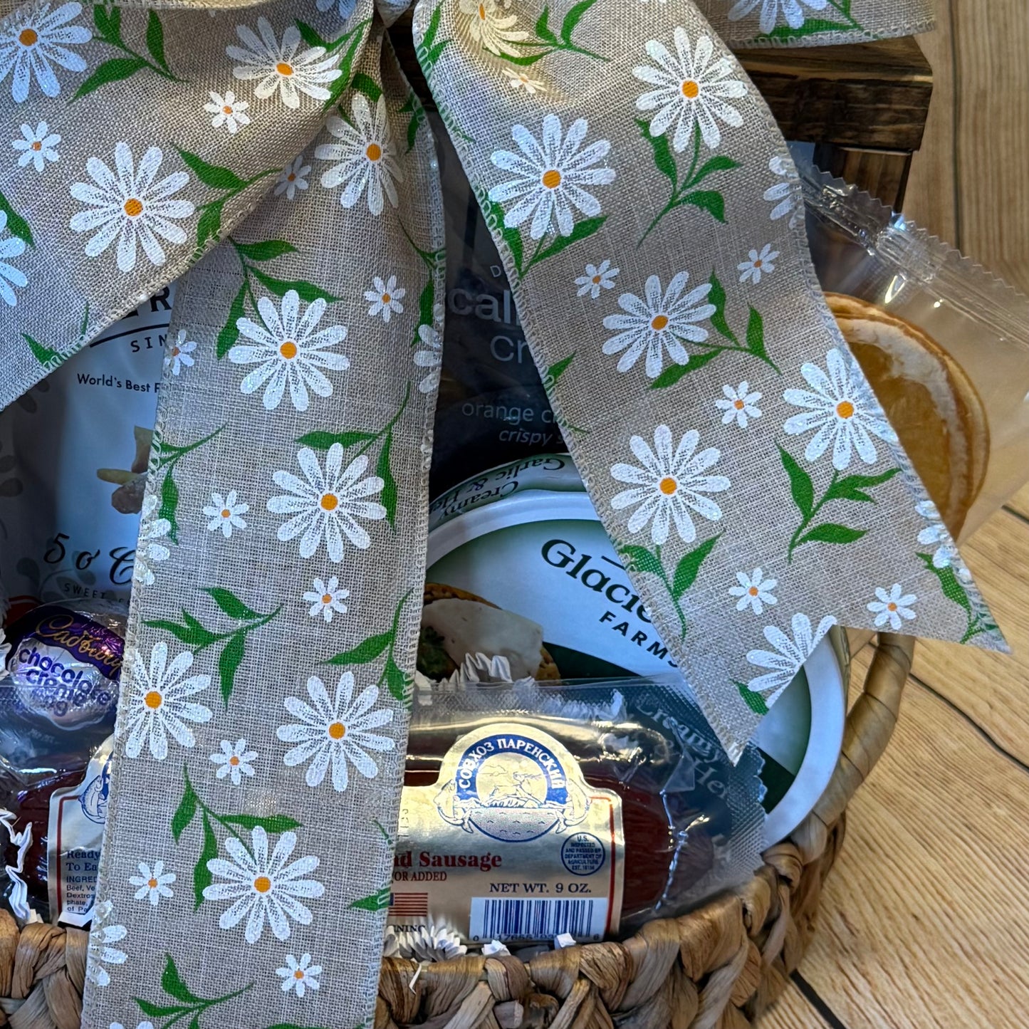 Easter Delights Gift Basket with assorted treats, including cream cheese, smoked sausage, Cadbury Egg, citrus crisps, cookie, and a bag of tea or coffee, adorned with a daisy burlap ribbon.