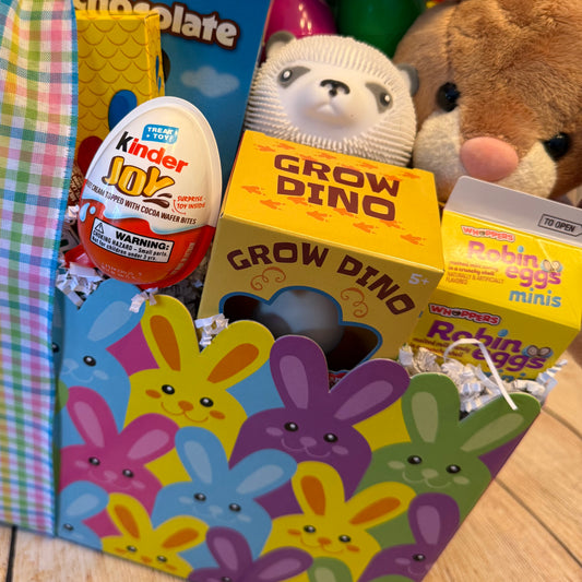 Easter Fun Gift Basket with a colorful bunny print, filled with Kinder Joy egg, Grow Dino kit, Robin Eggs candy, silicone toy, plush animal, and plastic eggs.