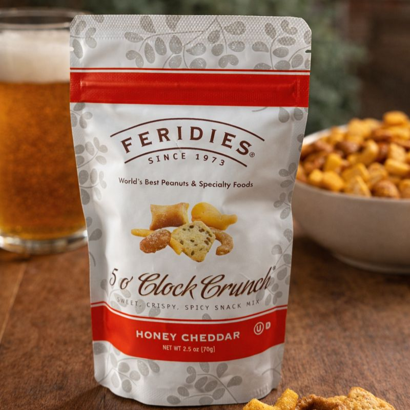 Feridies 5 o'Clock Crunch Honey Cheddar snack mix packaging on a wooden surface with a glass of beer and bowl of snacks in the background.