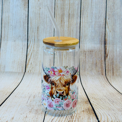 A clear glass tumbler with a floral pattern consisting of a Highland cow, pink flowers and green leaves, including a bamboo lid and a glass straw.
