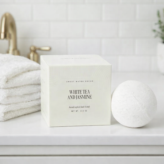 White Tea & Jasmine Bath Bomb on a bathroom countertop, accompanied by a stack of white towels and a brass faucet, offering a luxurious, non-toxic, vegan soak.