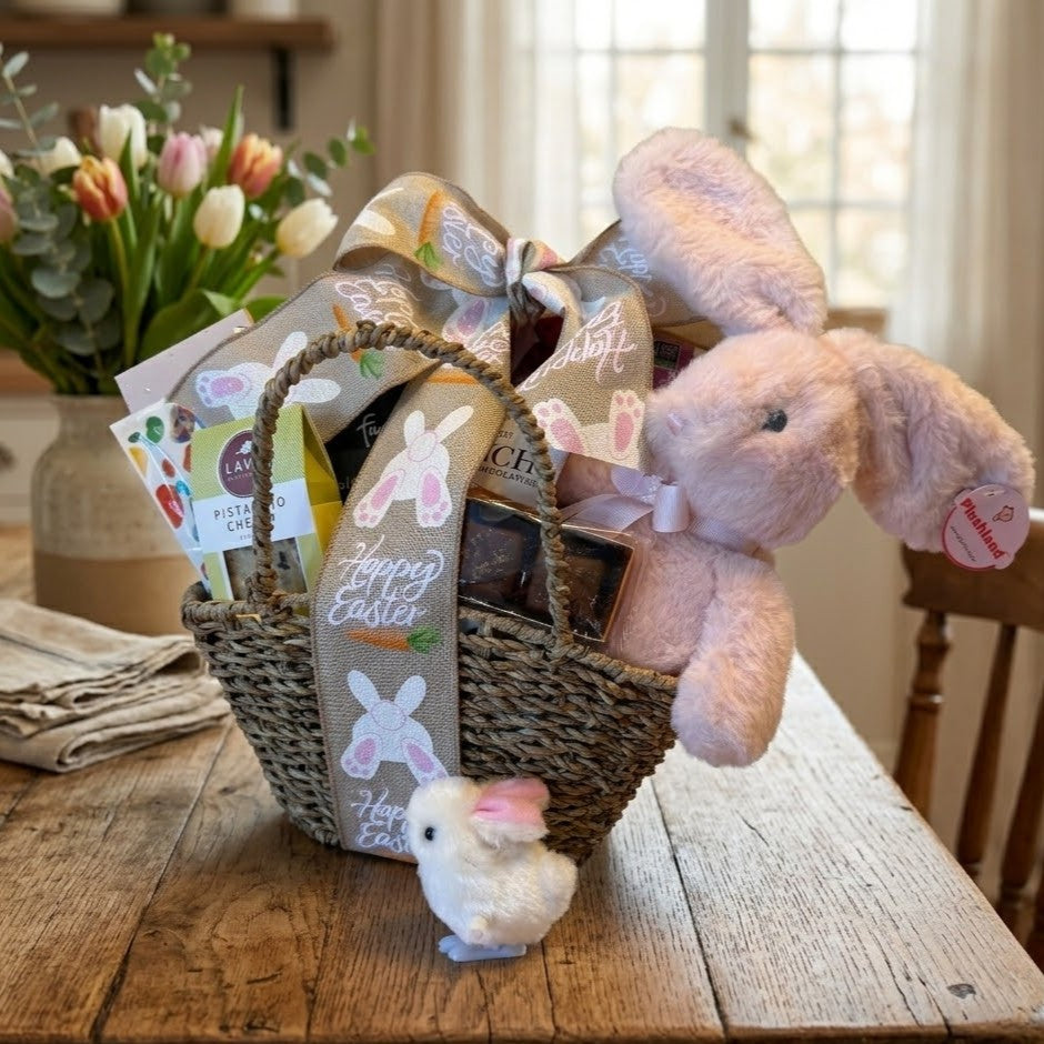The Little Bunny Easter Gift Basket features a plush pink rabbit, Happy Easter burlap ribbon, assorted confections, and a decorative bunny on a rustic table.