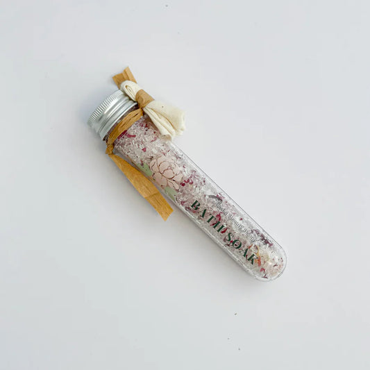 A clear glass jar with a label reading 'BATH SOAK', filled with pink and white bath salts. The jar is sealed with a silver lid and has a beige ribbon tied around it.