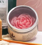 A clear container with a red peony candle inside, set on a wooden surface with a book and a candle holder in the background.