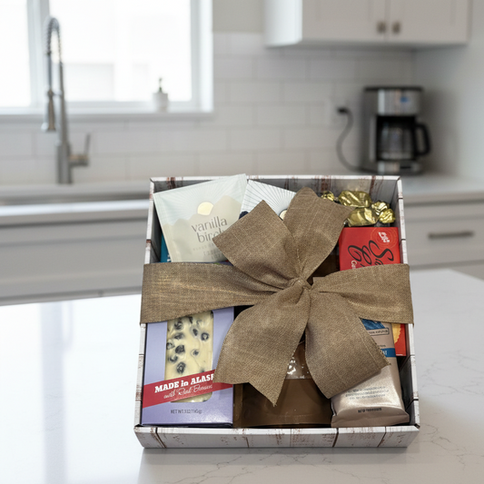 Susitna Alaskan Gift Crate on countertop, filled with assorted treats like Vanilla Birch tea, Alaskan chocolate, gold-wrapped candies, all in a burlap-tied container.