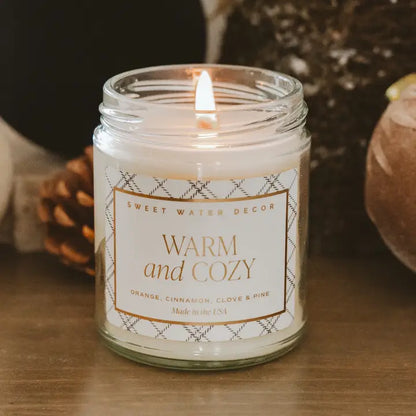 Candle in a glass jar with a label reading 'Warm and Cozy' on a wooden surface.