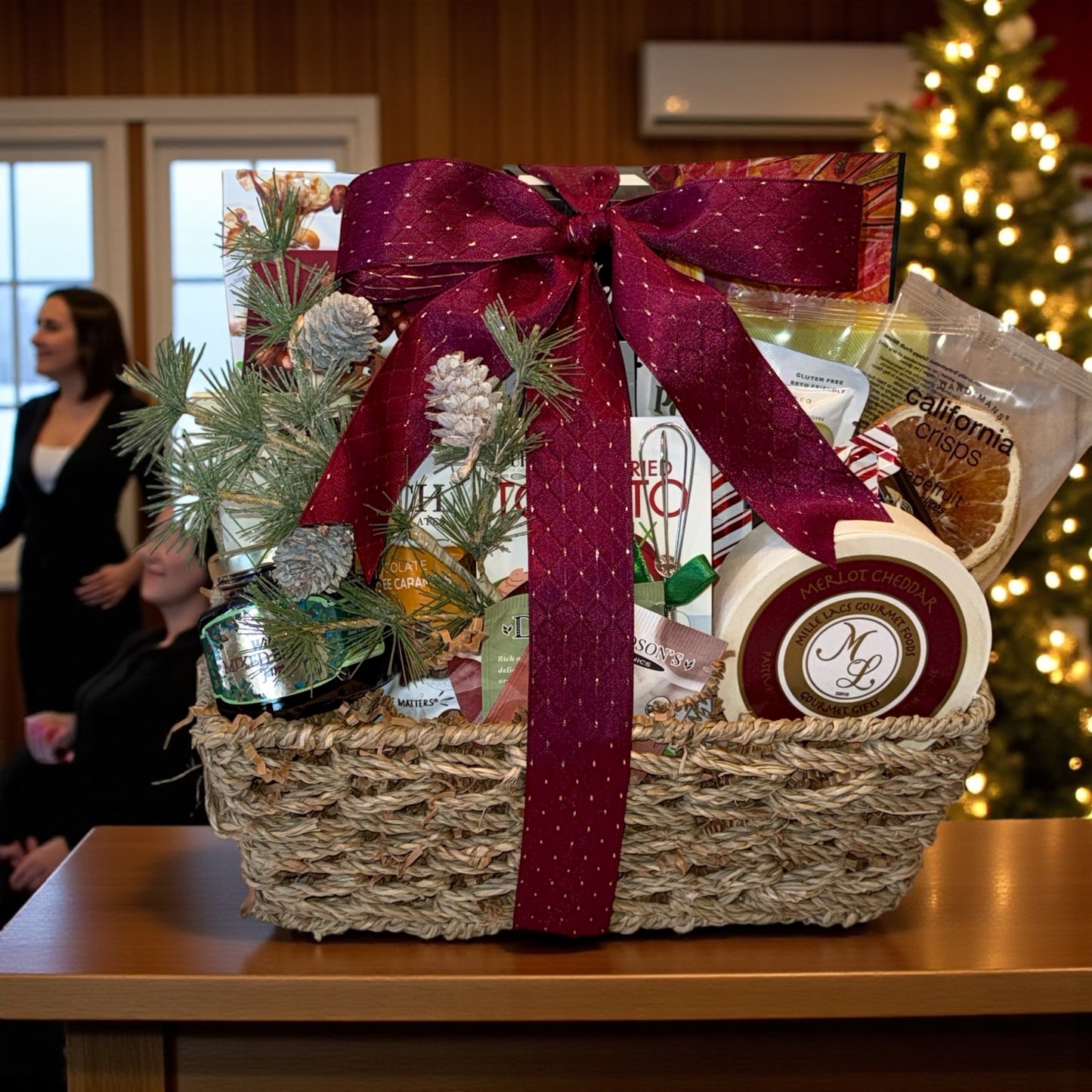 Gift basket with a red ribbon on a table in front of a Christmas tree.