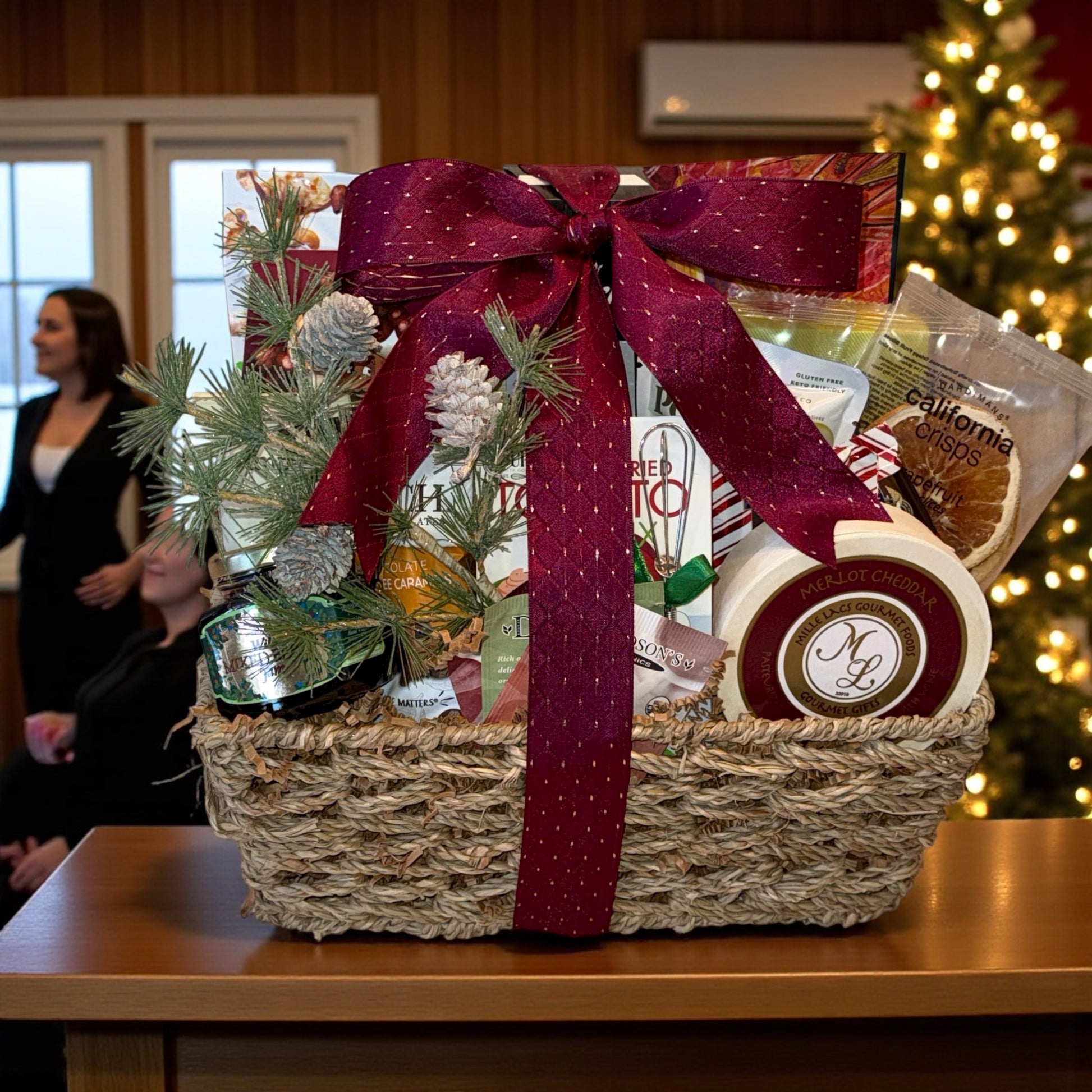 Gift basket with a red ribbon on a table in front of a Christmas tree.