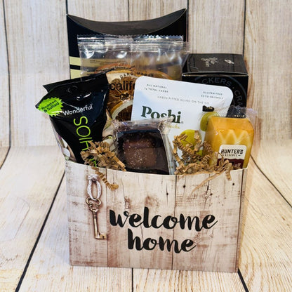 A gift basket with a 'welcome home' label, containing gourmet food items such as smoked salmon, artisan crackers, cheese, olives, pistachios, coffee, dried fruits, and chocolates, all packaged in a decorative box with a ribbon.