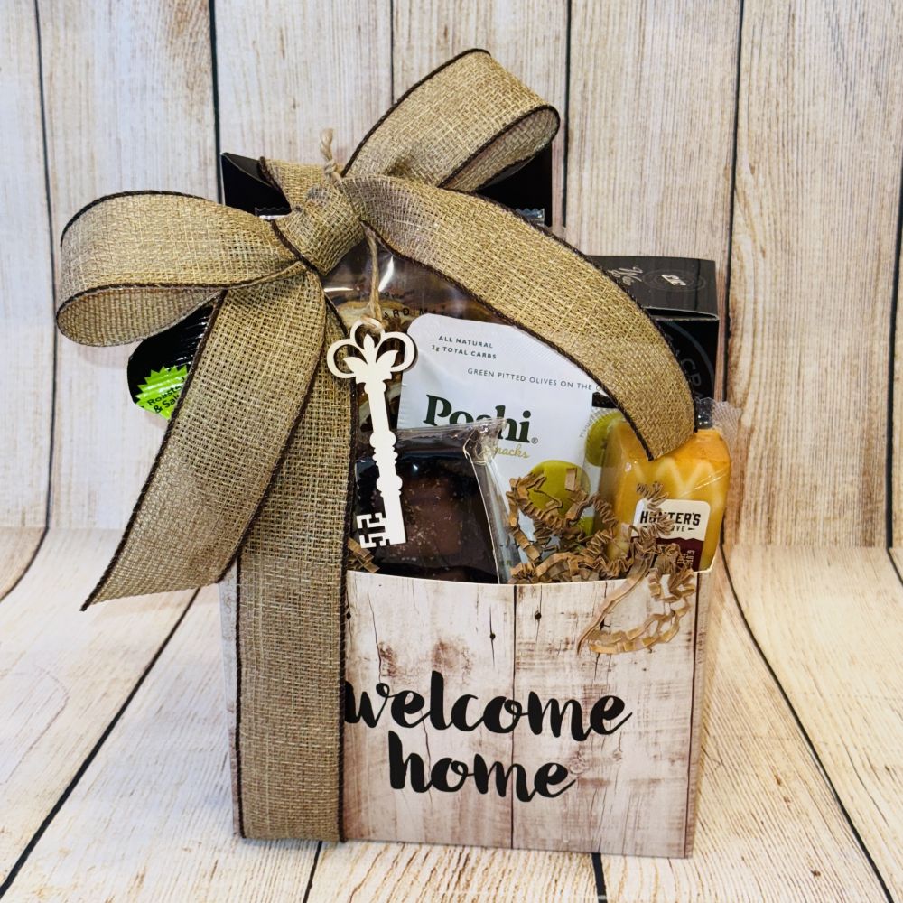 A gift basket with a 'welcome home' label, containing gourmet food items such as smoked salmon, artisan crackers, cheese, olives, pistachios, coffee, dried fruits, and chocolates, all packaged in a decorative box with a ribbon.